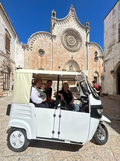 A white three-wheeled vehicle with passengers, parked in front of a historic building with intricate architecture. 3