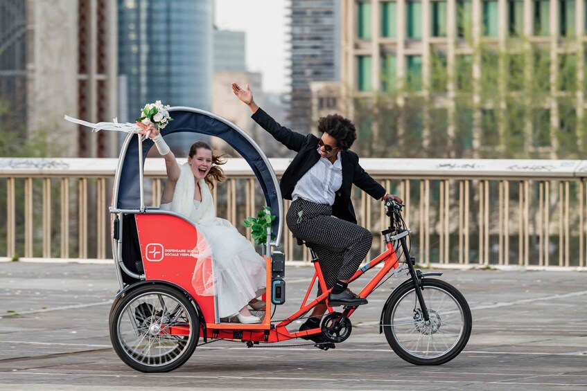 A wedding couple on a tandem bicycle with a canopy, in an urban setting with buildings in the background. 3