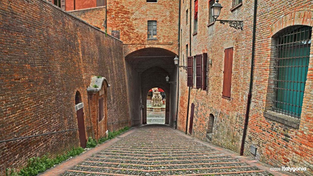A narrow alley with cobblestone path, brick buildings, and a central archway leading to a fountain. 3