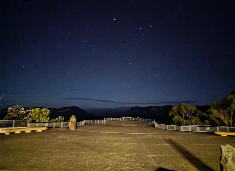 A nighttime view of a paved area with a railing, a large rock, and a clear sky with stars. 3