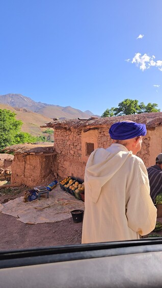 A man in traditional attire standing in front of a rustic building with a mountainous backdrop. 1