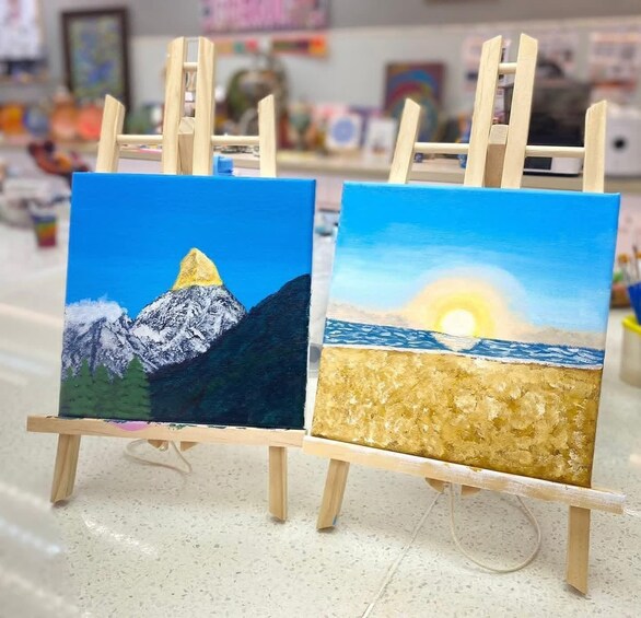 Two paintings on easels depicting mountains and a sunset over water. 2