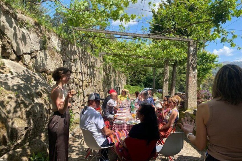 A group of people gathered around a table outdoors, possibly for a meal or event, with a stone wall and greenery in the background. 1