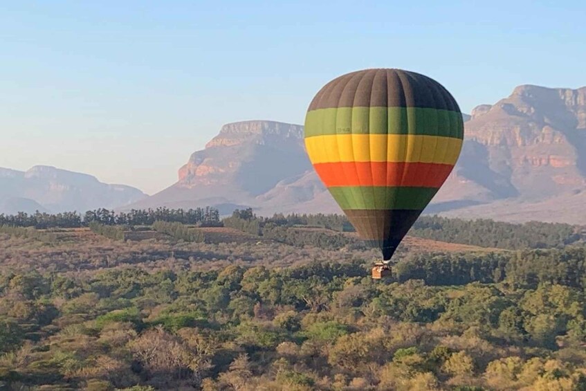 A hot air balloon with a multicolored pattern flying over a forested landscape with mountains in the background. 1