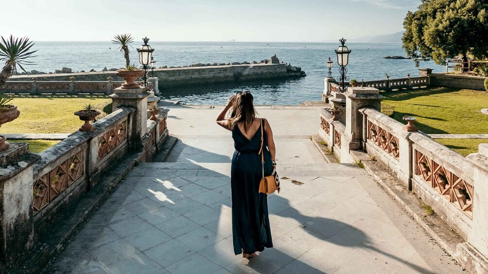 A woman walking down a stone pathway with ornate railings, overlooking a body of water and a distant ship. 1