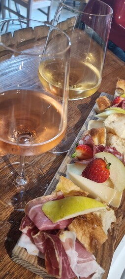 A wooden table with two glasses of white wine and a charcuterie board with meats, cheese, and fruits. 4