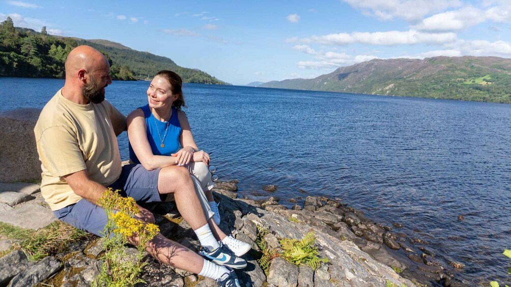 Two people sitting on a rocky shore by a lake with mountains in the background. 4