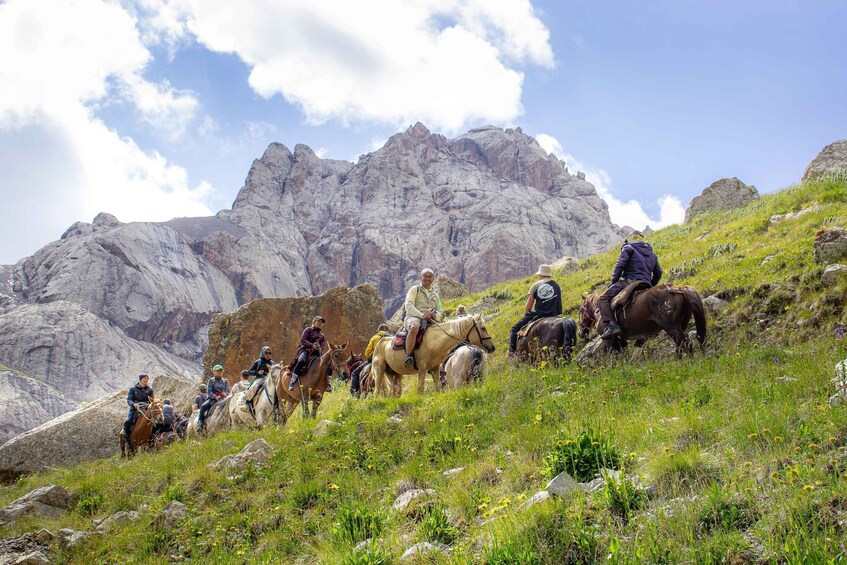 A group of people riding horses on a grassy hillside with rocky mountains in the background. 5