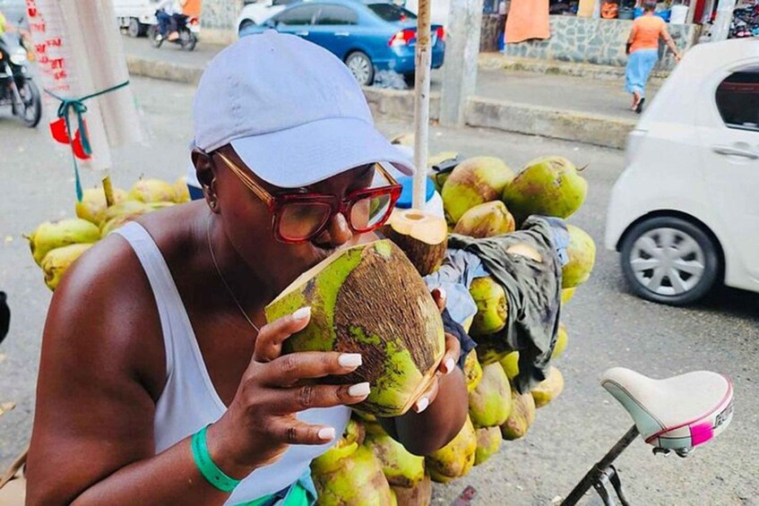 A person holding a coconut, surrounded by other coconuts, with a bicycle and cars in the background. 4