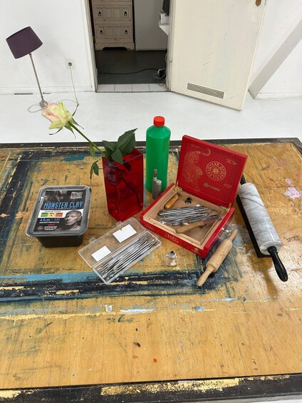 A table with crafting supplies including clay, a rolling pin, a vase with a rose, and a red box with tools. 2