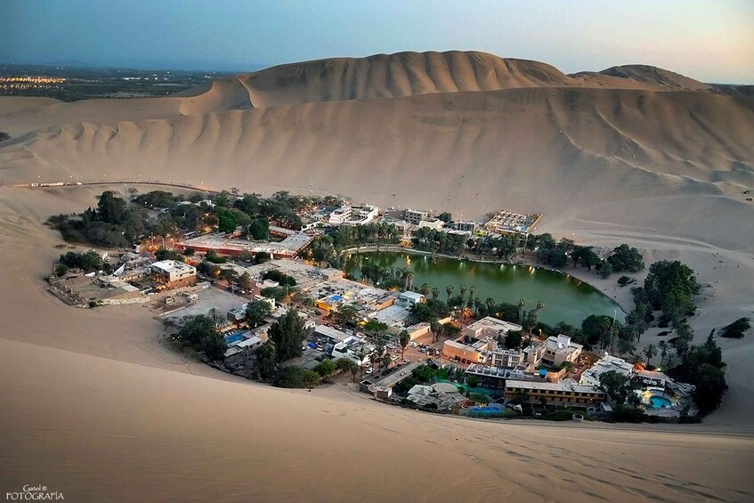 A desert landscape with sand dunes and a small settlement with buildings and a body of water. 5