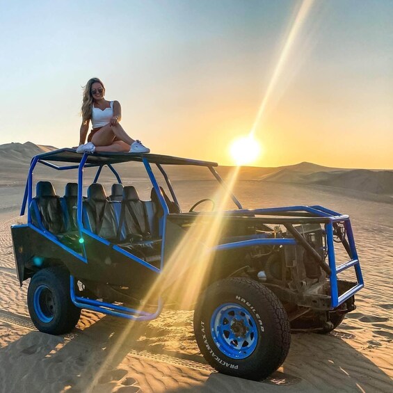 A dune buggy parked on sand dunes with a person sitting on the roof, sun setting in the background. 1