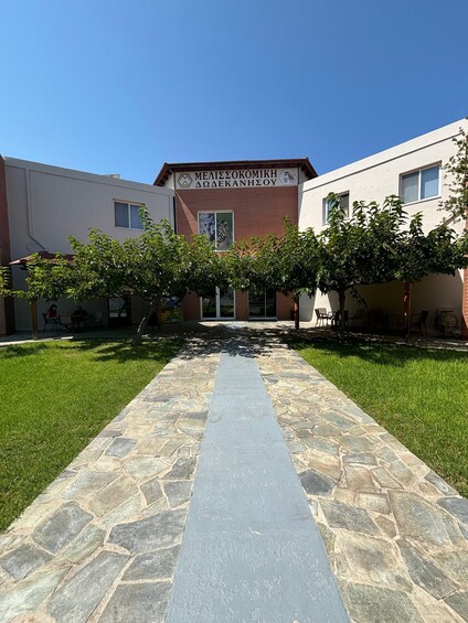 A building with a sign reading 'MEALIESSEOKOMIKH ΔΩΛΕΚΑΝΗΣΟY' and a stone pathway leading to it. 5