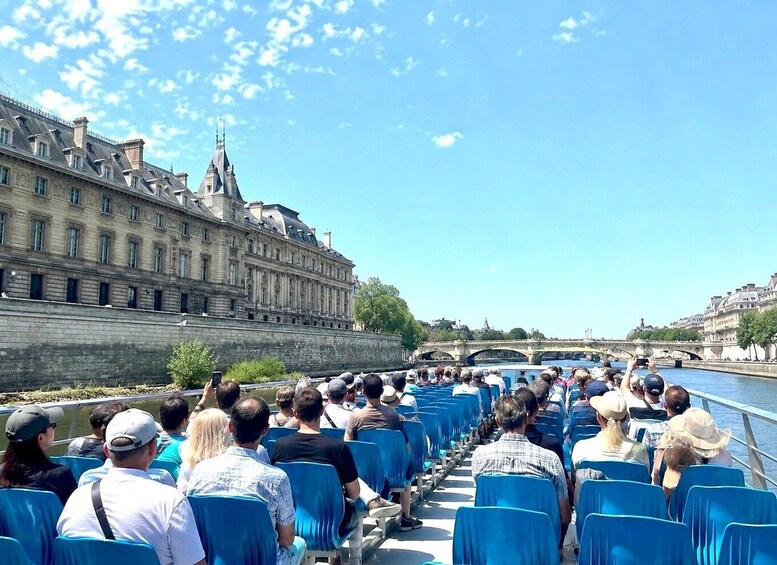 A river cruise with passengers seated in rows, a historic building on the left, and a bridge in the background. 3