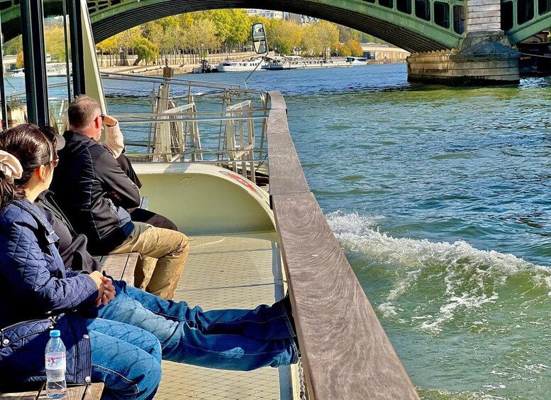 People sitting on a boat under a bridge, with a river and boats in the background. 4