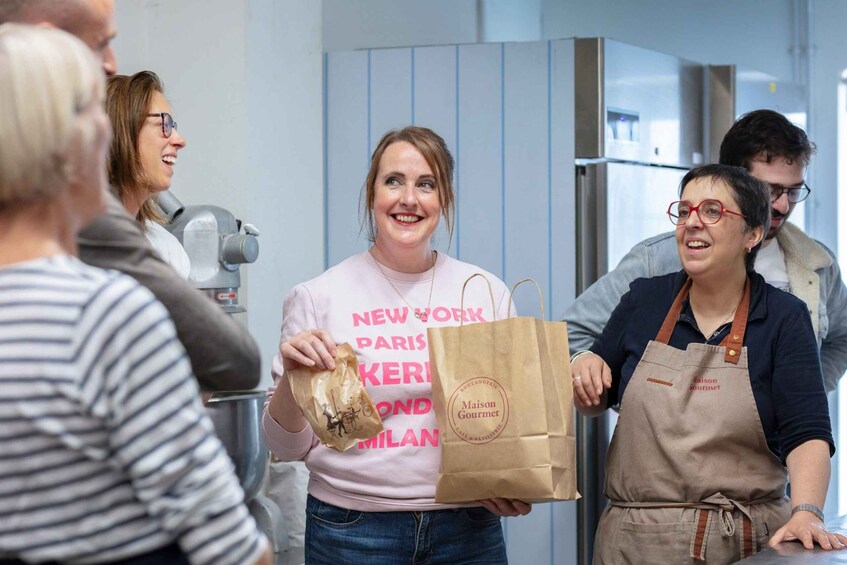 A person holding a brown paper bag with 'Maison Gourmet' printed on it, standing in a kitchen with stainless steel appliances. 5
