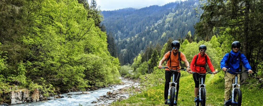 Three cyclists on a mountain trail surrounded by dense forest and a flowing river. 1