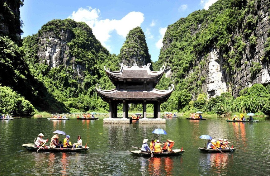 A traditional Chinese pavilion surrounded by lush greenery and rocky cliffs, with people paddling in boats on a calm river. 2