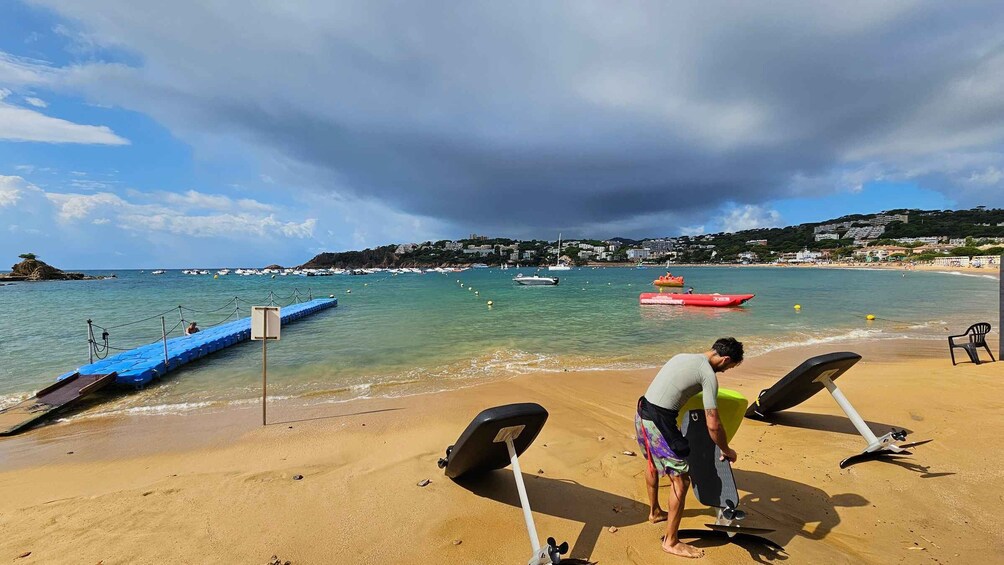 A beach with a man preparing a surfboard, a jet ski, and a row of boats in the water. 3