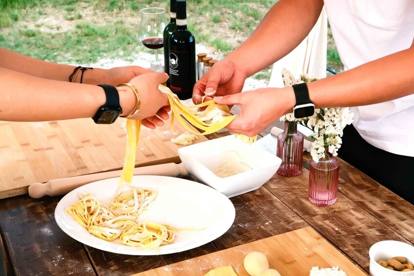 People preparing pasta on an outdoor table with a bottle of wine, flowers, and a rolling pin. 1