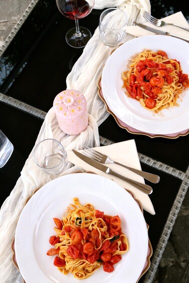 Two plates of pasta with cherry tomatoes on a table with a glass of red wine and a pink container. 4