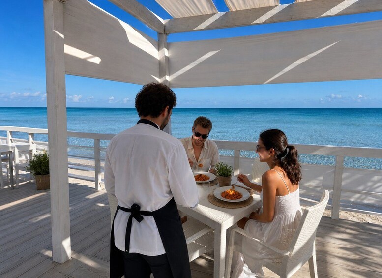 A couple dining at a white wooden table on a beach deck with a view of the ocean. 1