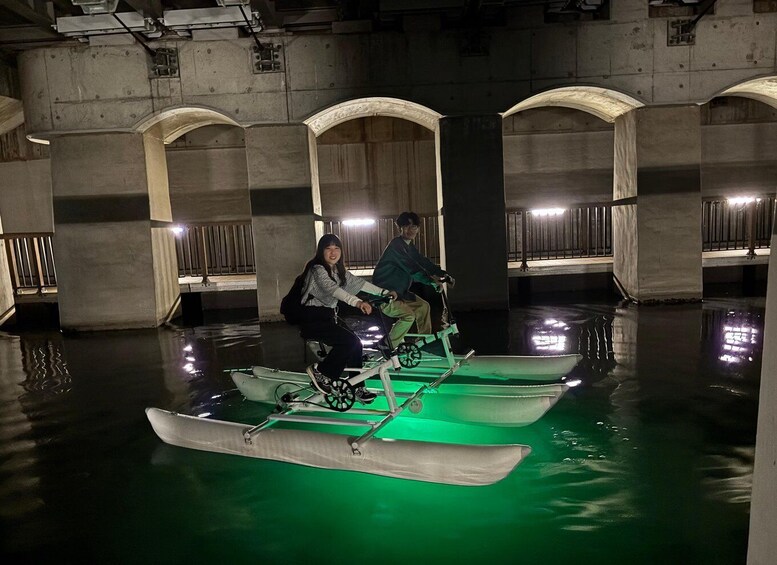 Two people on a green-lit boat in an indoor waterway with concrete arches. 3