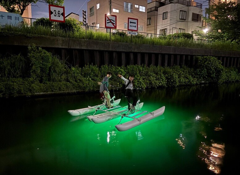 Two people on a green-lit boat in a canal with buildings in the background. 5
