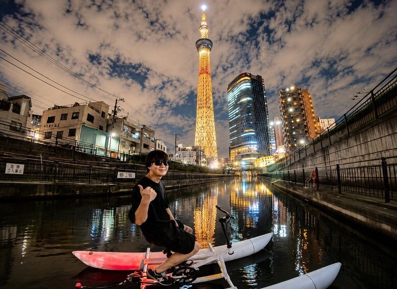 A nighttime cityscape with a lit tower, a river reflecting lights, and a person on a kayak. 1