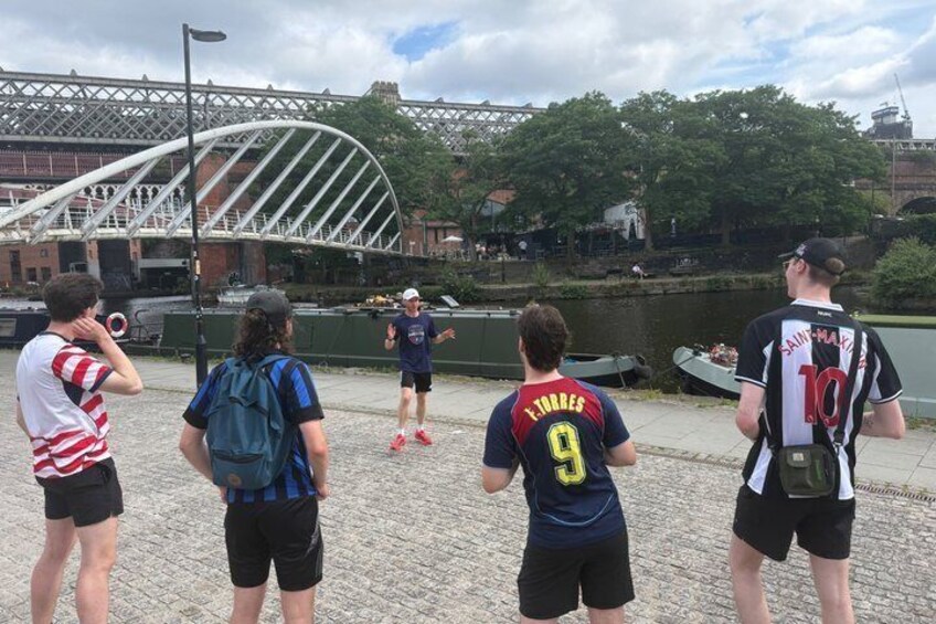 A group of people standing on a cobblestone path by a river, with a bridge and trees in the background. 4