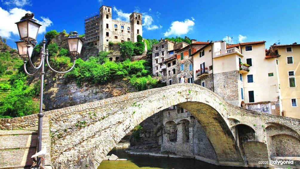A stone bridge with arches, a street lamp, and buildings on a hillside. 1