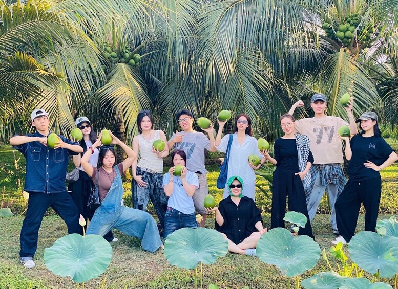 A group of people holding coconuts in a tropical setting with palm trees and large green leaves in the background. 2