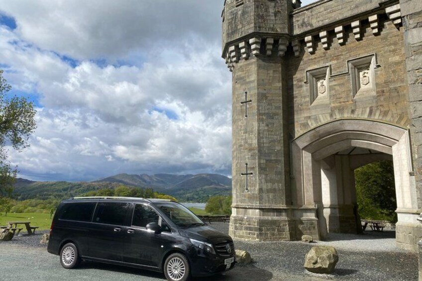 A black car parked in front of a stone building with a cross, a picnic table, and a mountainous landscape in the background. 1