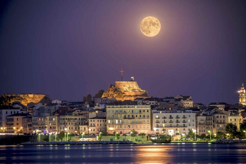 A cityscape at night with a prominent hilltop fortress, a full moon in the sky, and illuminated buildings along the waterfront. 1
