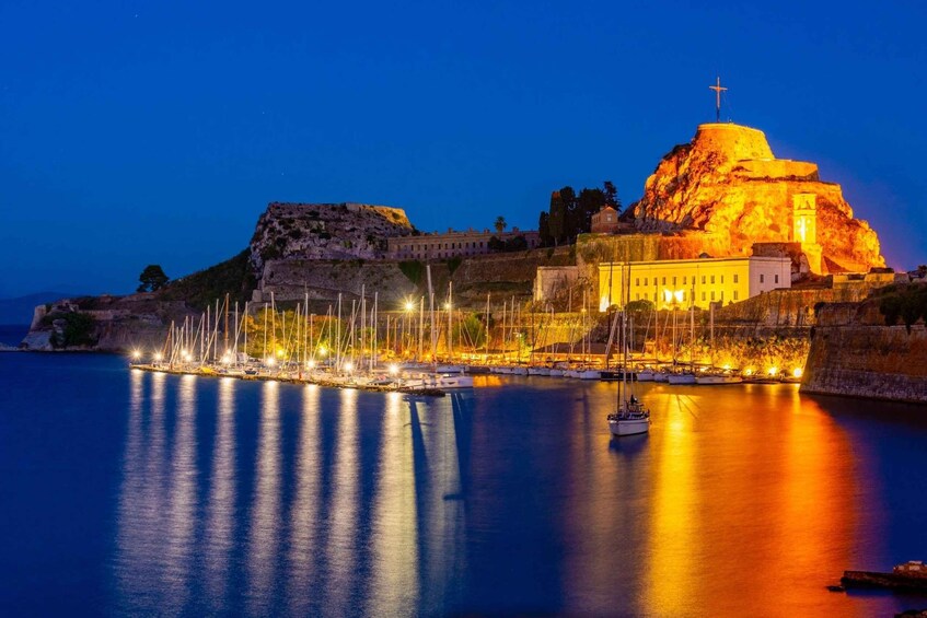 A coastal town at night with illuminated buildings, a prominent hilltop structure, and a marina with boats. 3