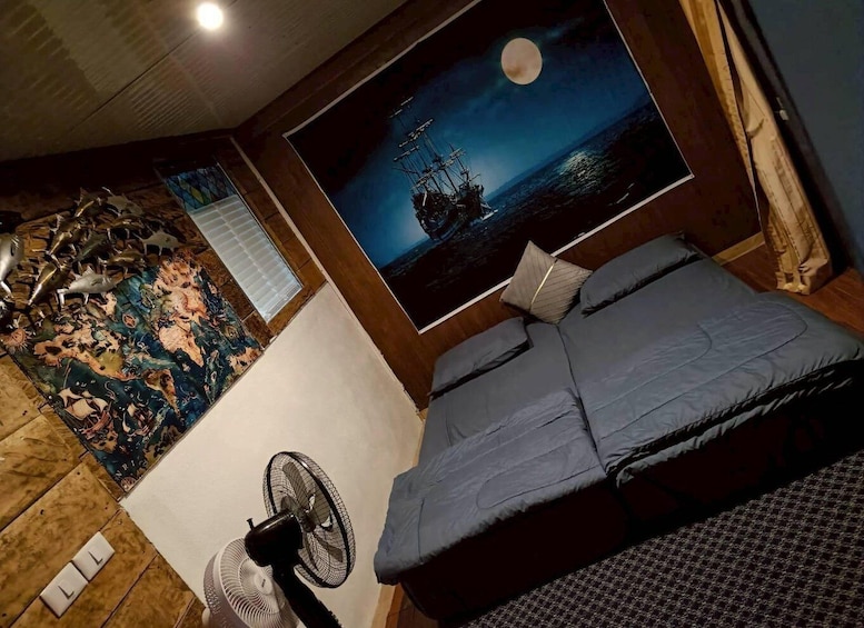 A bedroom with a bed, a fan, a painting of a ship at sea, and a decorative rug. 4