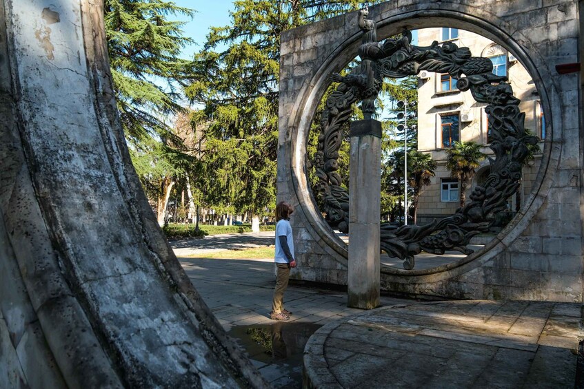A person standing in front of a circular stone structure with intricate carvings, surrounded by trees and a building in the background. 3