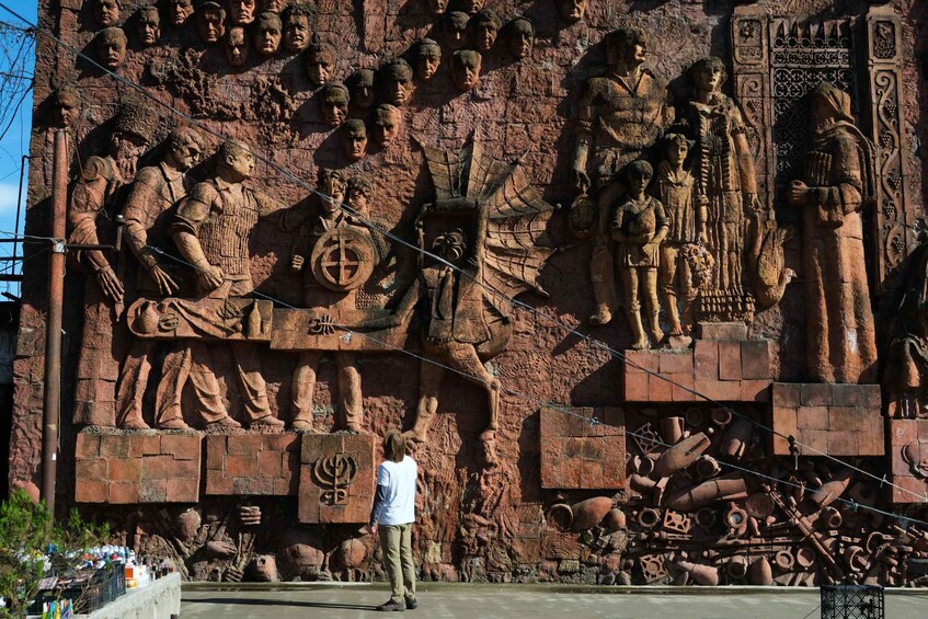 A large stone relief with figures and symbols, a person standing in front, and a pile of metal objects at the base. 2
