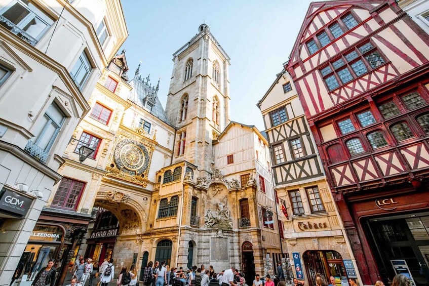 A historic European square with ornate architecture, including a clock tower and a building with a facade featuring a mix of white and red timber framing. 1