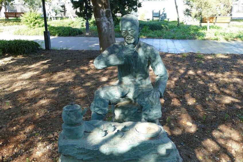 A statue of a seated man with a contemplative expression, surrounded by a landscaped park with trees and a bench in the background. 2