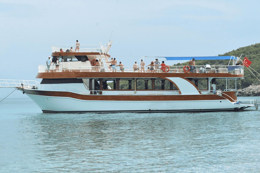 A white and brown boat with people on deck, docked near a shoreline with a flag. 4
