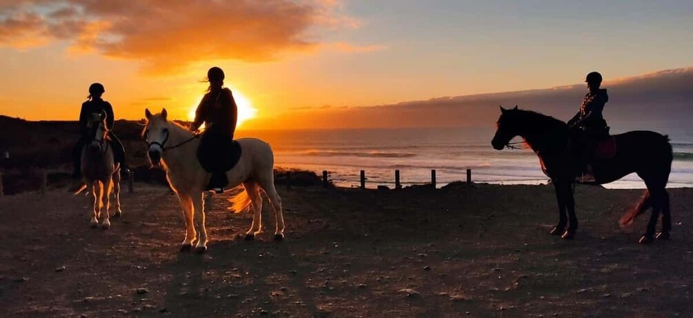 Three riders on horses along a coastal path during sunset. 1