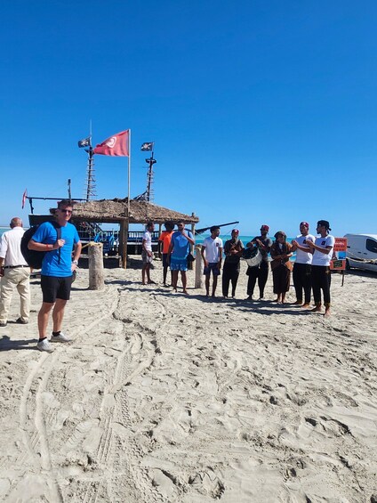 A group of people on a sandy beach with a thatched-roof structure and flags in the background. 3