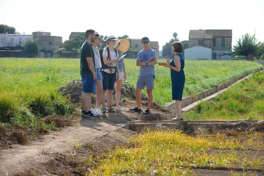 A group of people standing on a dirt path in a field with grass and a few buildings in the background. 2