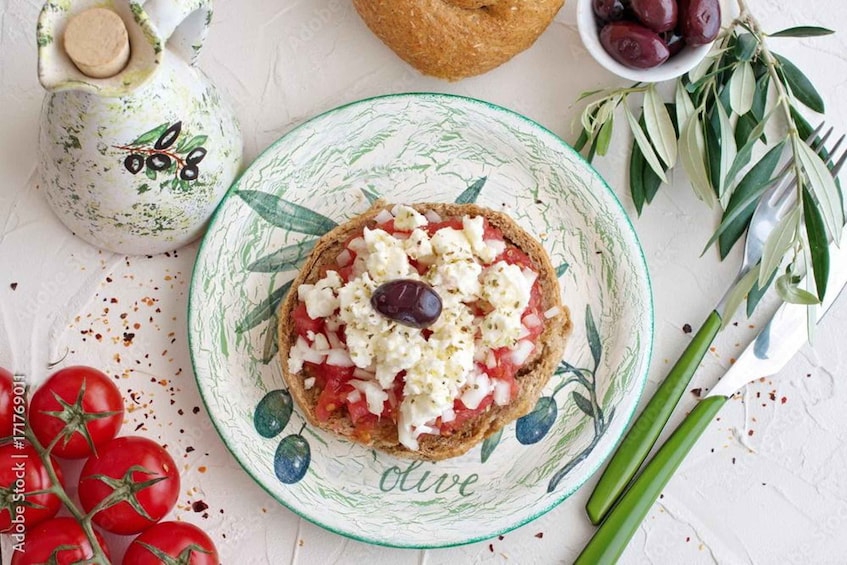 A plate with a slice of bread topped with tomatoes, feta cheese, and an olive. There are tomatoes, a bowl of olives, a loaf of bread, and green herbs in the background. 2