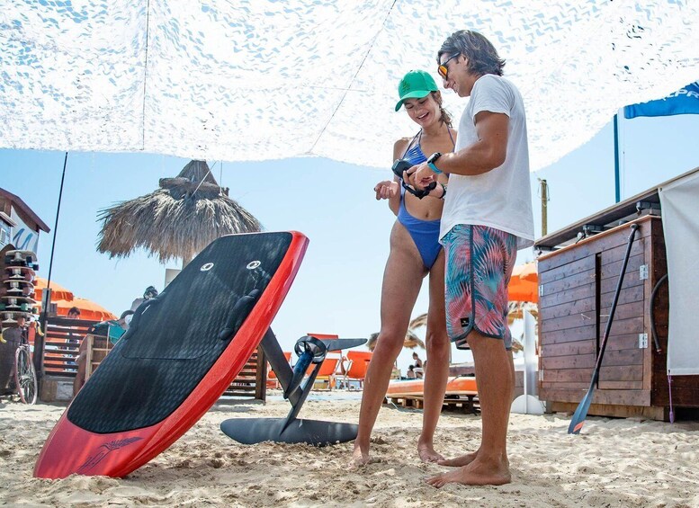 A couple on a beach with a surfboard, umbrellas, and a thatched structure. 2