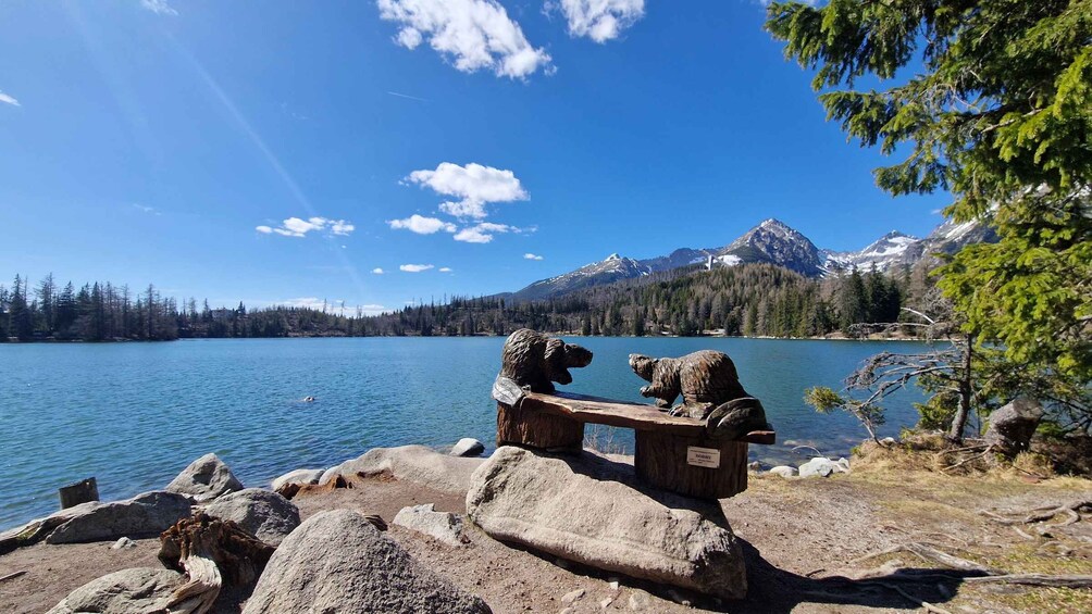 A lake with a wooden bench and two stone sculptures, surrounded by mountains and trees. 4