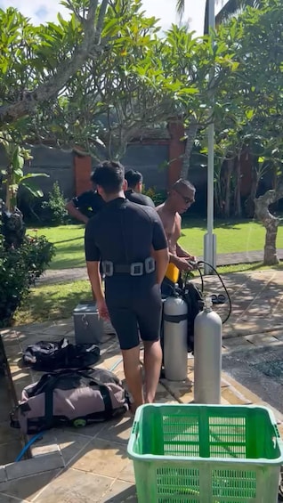 A person in a wetsuit preparing scuba gear on a patio with a green basket and a bag nearby. 3