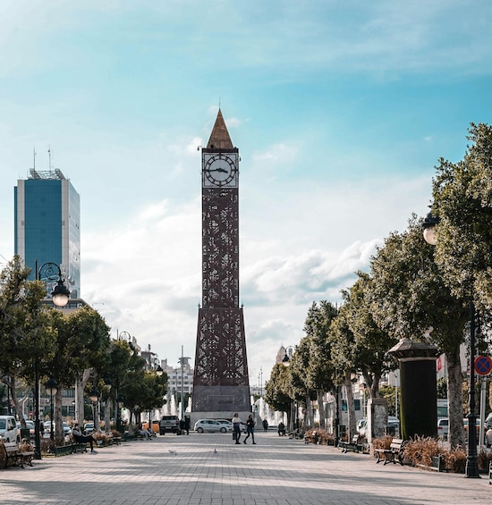 A tall tower with a clock, surrounded by trees and benches in a city square. 1