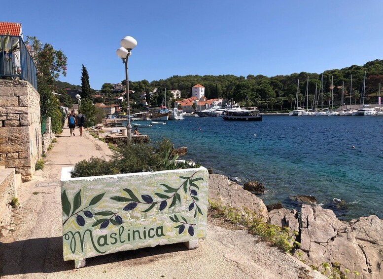 A coastal town with a sign reading 'Maslinica,' boats docked along the harbor, and a clear blue sky. 3